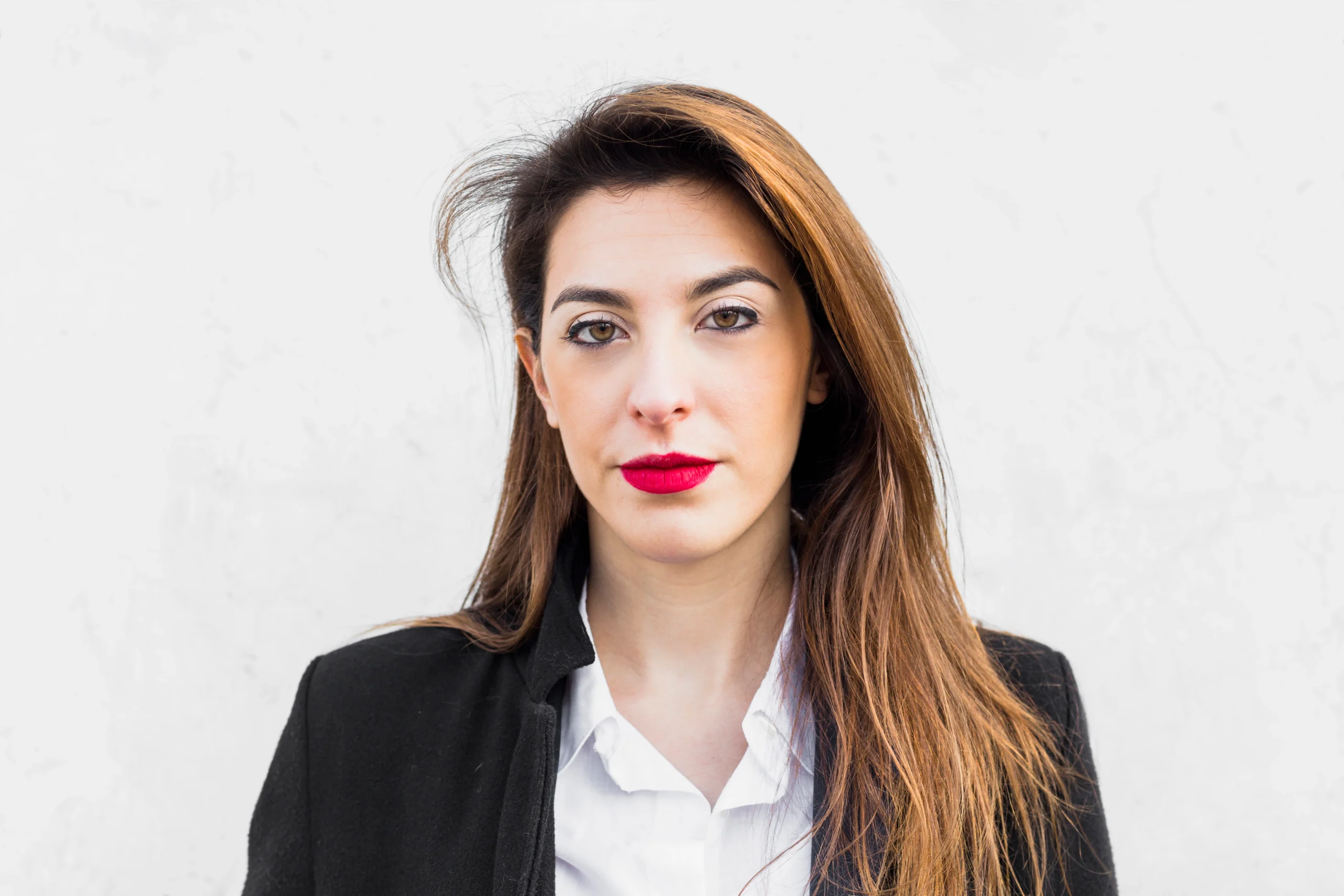 Professional headshot of a woman with long brown hair and red lipstick, wearing a white shirt and black blazer, against a light-textured neutral background.