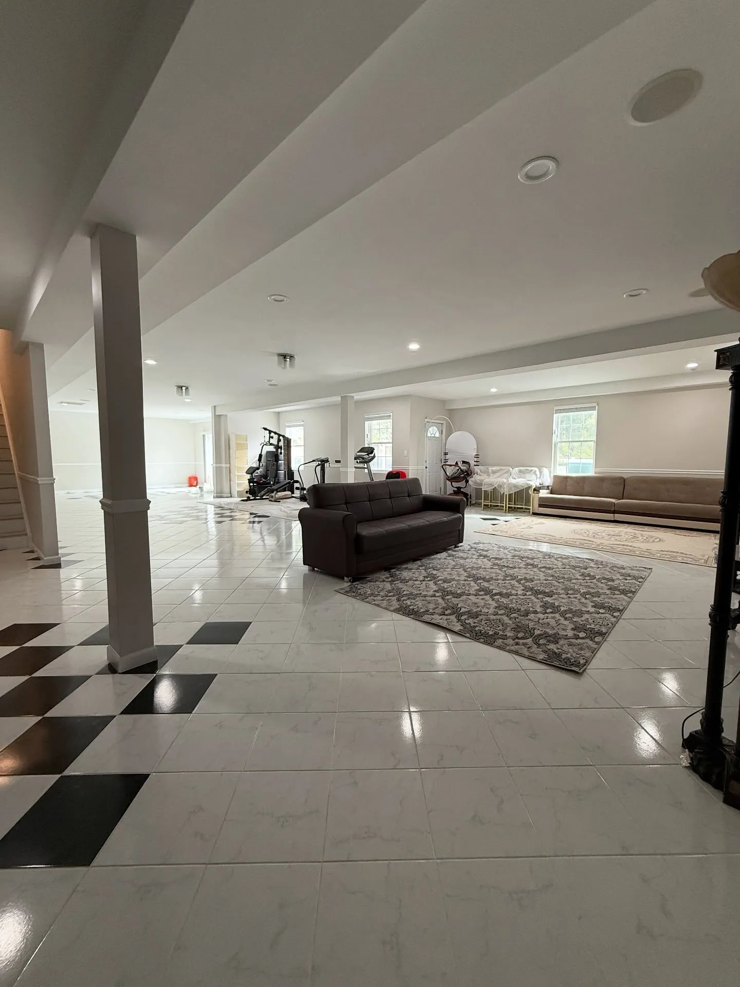 A large, brightly lit finished basement renovation by Dream Space Remodeling featuring polished white tile flooring with a checkered accent, recessed ceiling lights, a home gym area, and comfortable seating zones.