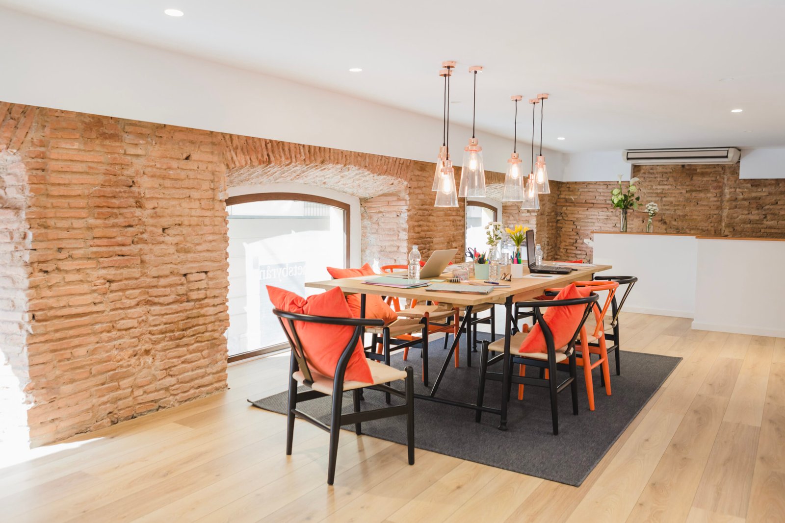 Modern office meeting space by Dream Space Remodeling featuring a long wooden conference table, orange and black chairs, exposed brick walls with arched windows, and elegant glass pendant lighting.