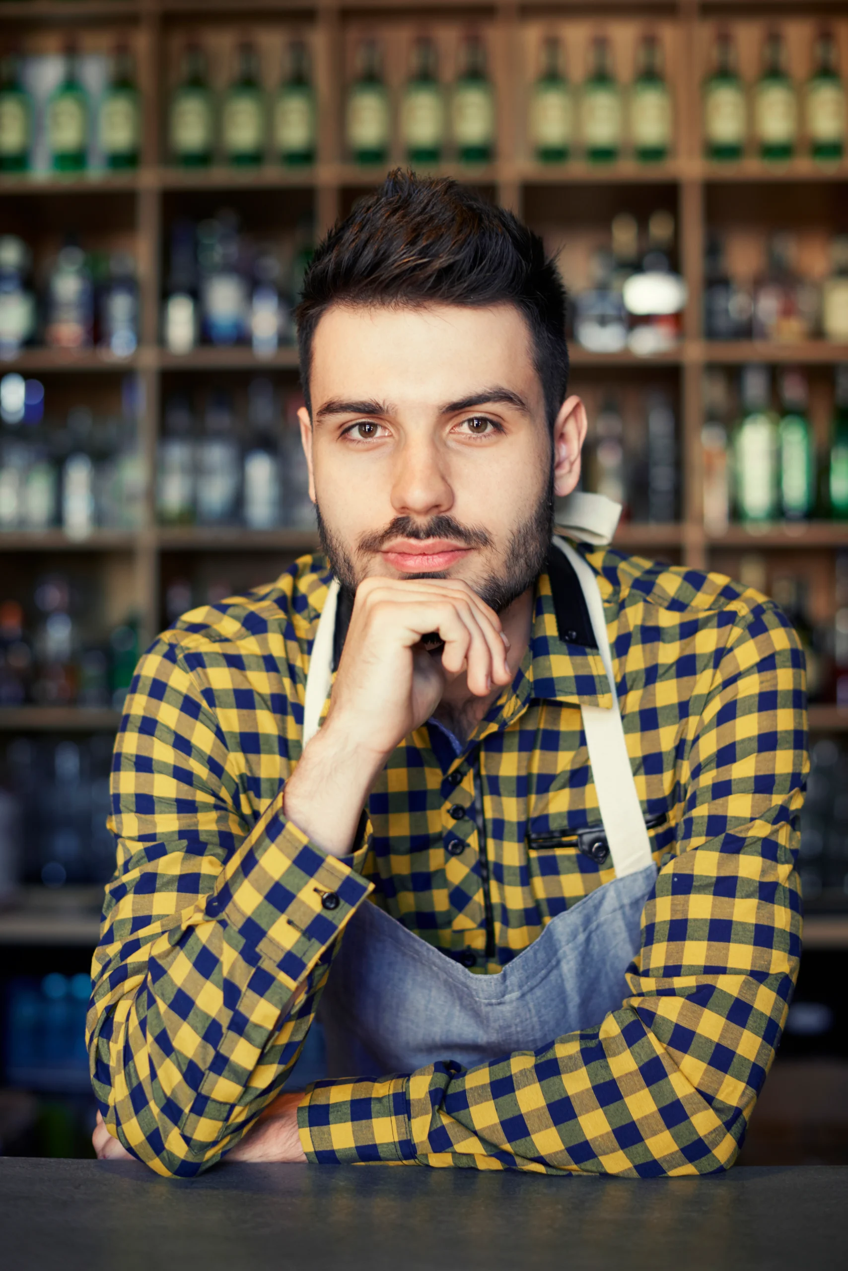 A young man with short dark hair and a light beard, wearing a green, white, and black plaid shirt, smiling and pointing his left hand to the side against a solid light gray background.