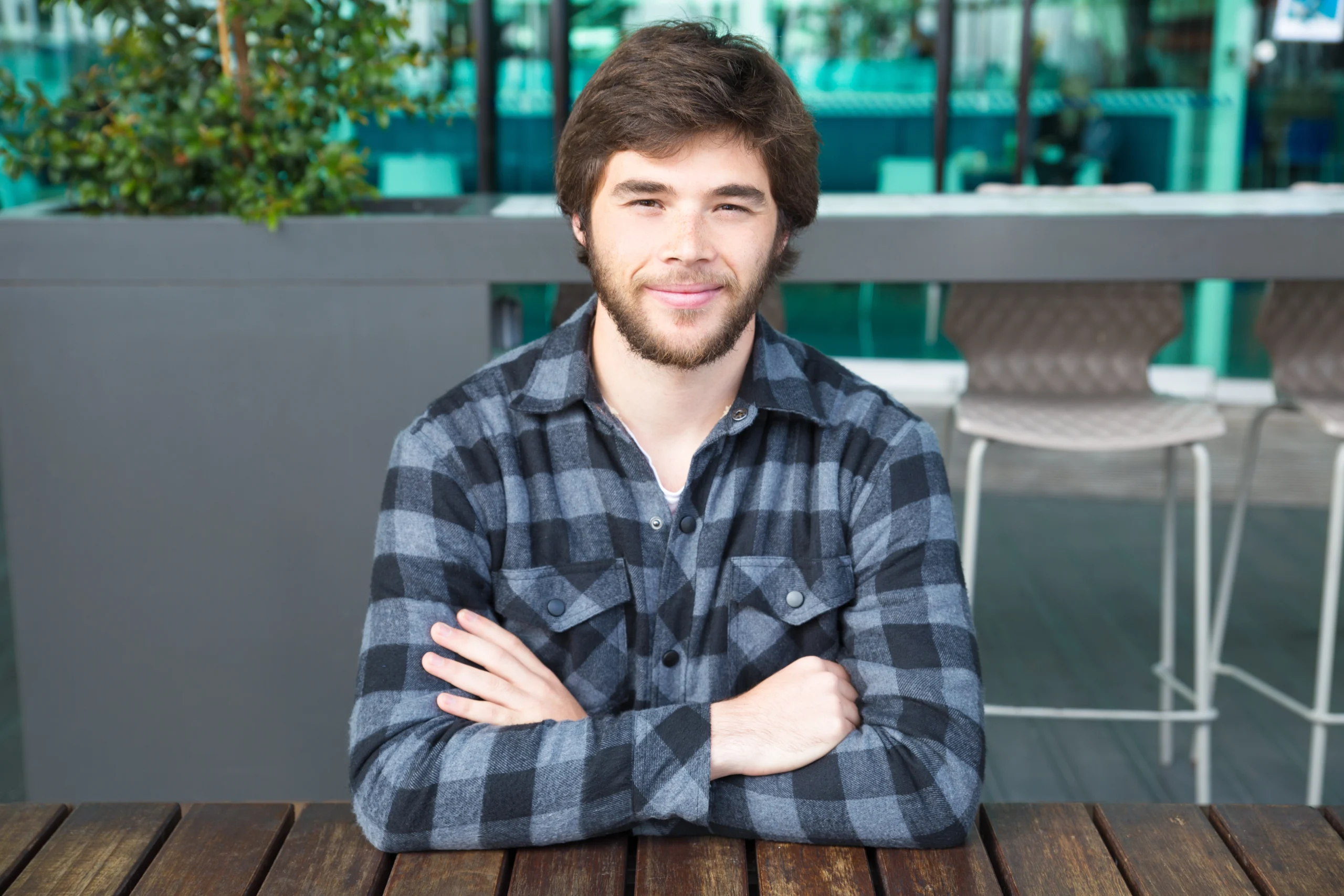 A smiling young man with a neat beard, wearing a gray and black plaid shirt over a white t-shirt, sitting at a dark outdoor table with his arms crossed.