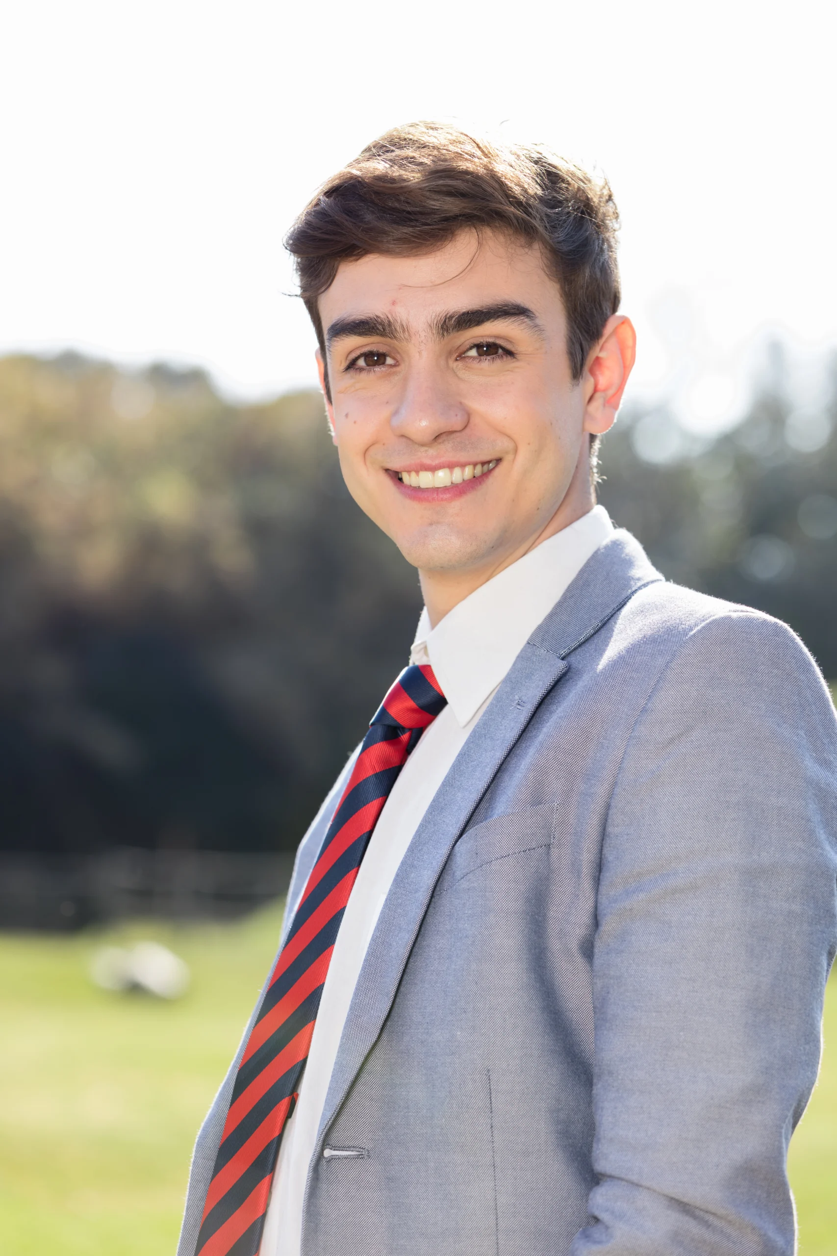 Professional outdoor headshot of a smiling young man wearing a light gray blazer, white shirt, and a red and navy striped tie against a blurred natural green background.