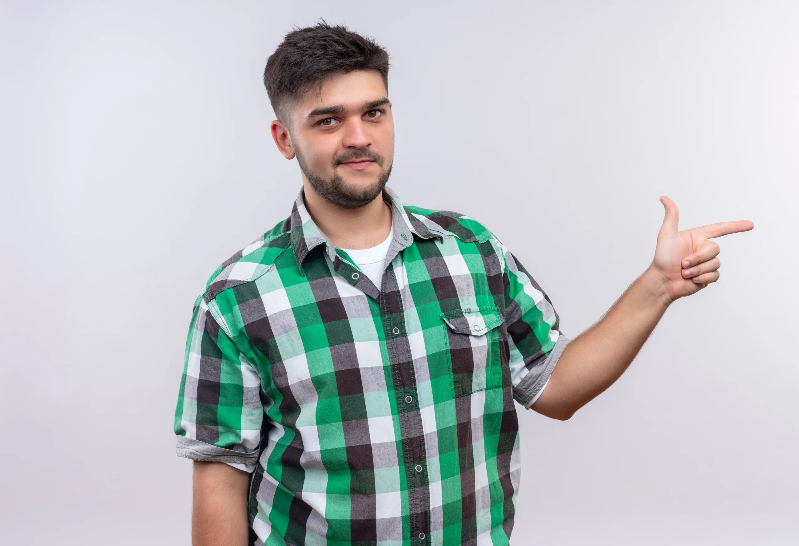 A smiling young man with a beard, wearing a green plaid shirt over a white t-shirt, pointing both hands upwards against a plain gray background.